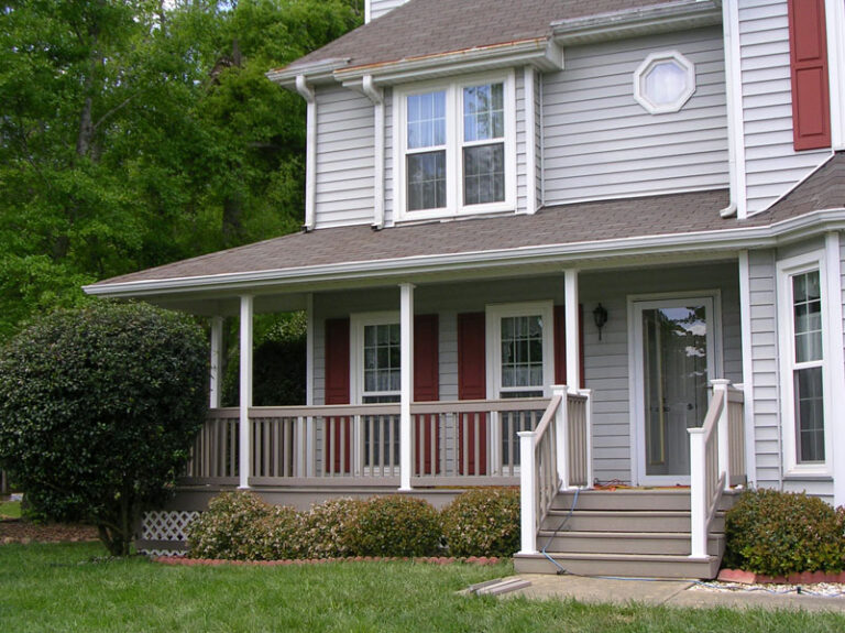 Two-story gray house with white trim, red shutters, and a front porch surrounded by greenery.