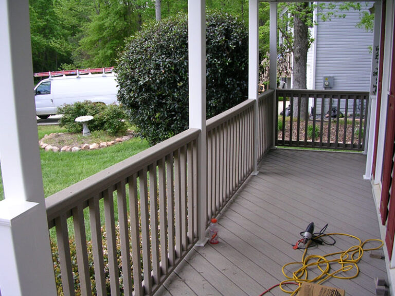 A view of a porch with beige decking, wooden railing, and green surrounding landscaping, featuring a white van parked in the background.