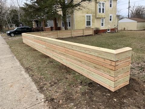 A newly constructed wooden planter box with horizontal slats, situated on a grassy area next to a sidewalk and a residential building.