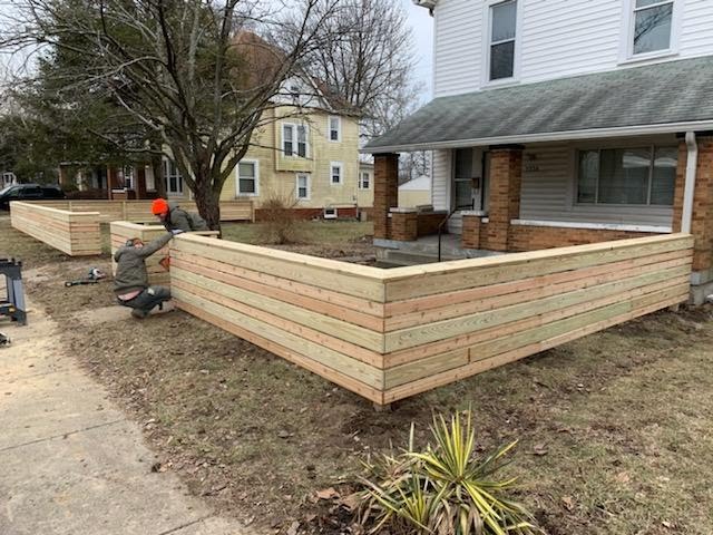 Two workers constructing wooden garden beds in a residential yard with a house in the background.