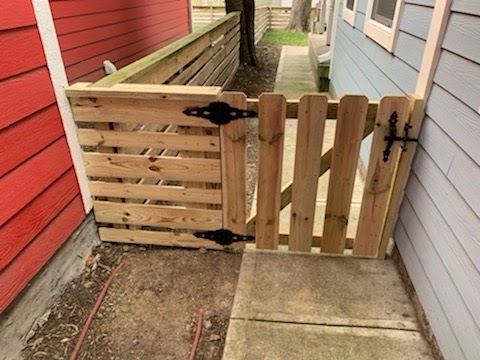 Wooden gate with decorative black hinges, set between two fences, leading to a pathway beside a house.