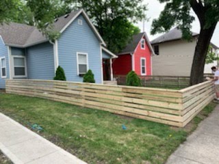 A new wooden fence surrounds a grassy yard in front of a blue house, with a red house visible in the background, creating a neat outdoor space.