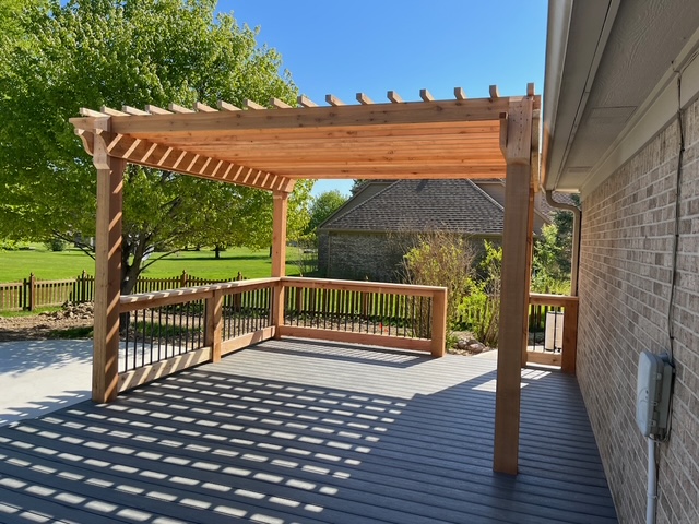 A wooden pergola shaded deck overlooking a grassy area and trees, with a clear blue sky above.