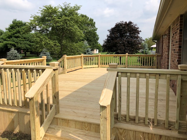 Newly constructed wooden deck with railing, surrounded by green trees and a grassy yard.