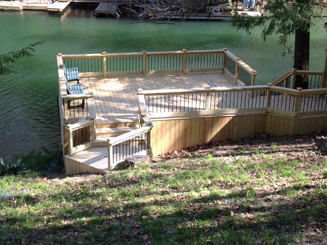 A wooden deck with a railing and two chairs overlooking a green lake, set in a grassy area.