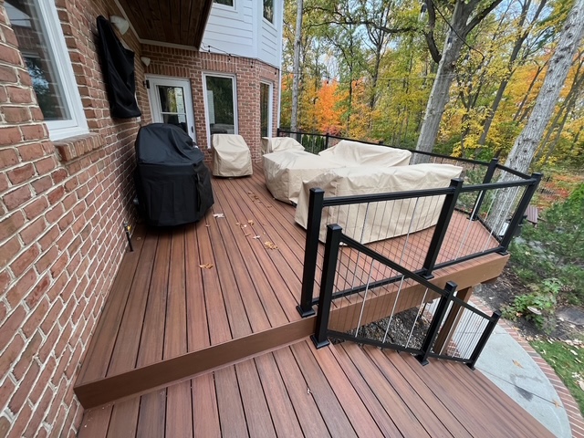 A modern wooden deck with patio furniture covered in beige fabric, showcasing a serene wooded backdrop with autumn foliage and a black railing.