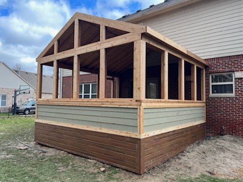 A newly constructed wooden porch with a gabled roof, featuring open sides and a combination of light green and dark brown siding, adjacent to a brick house.