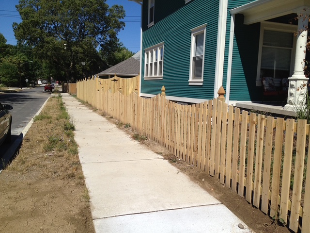 View of a newly constructed wooden picket fence along a sidewalk next to a blue house on a sunny day.