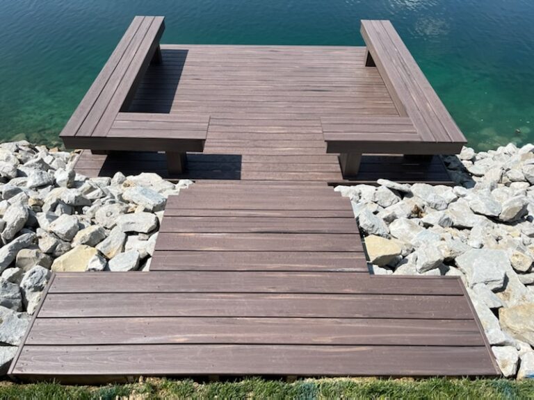 A wooden dock with two benches extends over calm, clear water, bordered by rocks and grass.