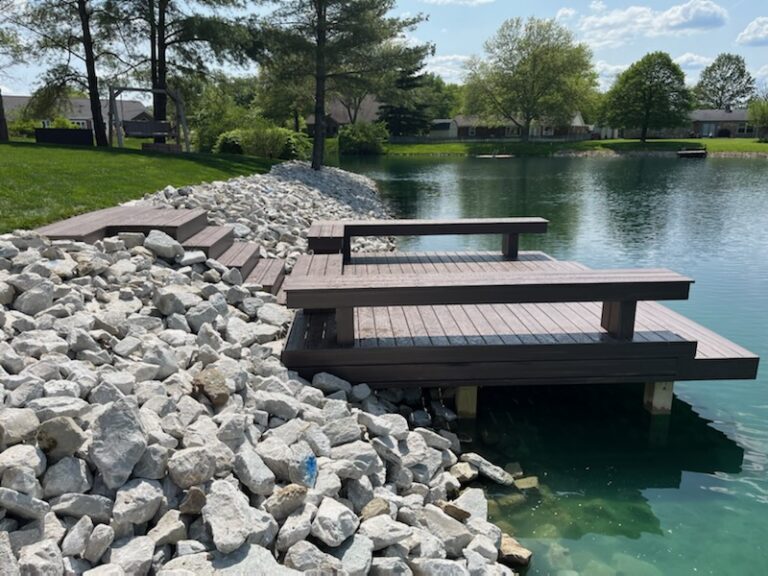 Wooden deck by a tranquil pond surrounded by rocks and lush green grass under a blue sky with fluffy clouds.