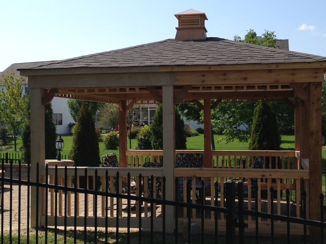 A wooden gazebo with a sloped roof, surrounded by greenery, featuring seating and a decorative lantern in a landscaped yard.
