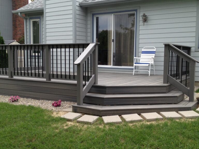 Deck with steps and a white and blue lawn chair, surrounded by grass and flowers near a light gray house.