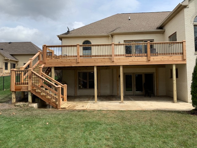 A newly constructed wooden deck with stairs, attached to a brick house, set against a grassy backyard under a cloudy sky.