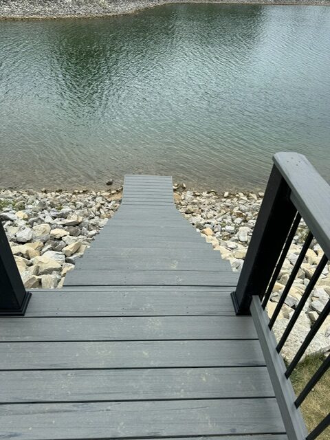 A wooden dock extends towards a calm body of water, bordered by rocks and greenery.