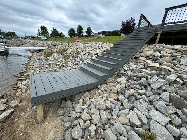 "Wooden staircase leading from a dock to a rocky shoreline by a lake, under cloudy skies."