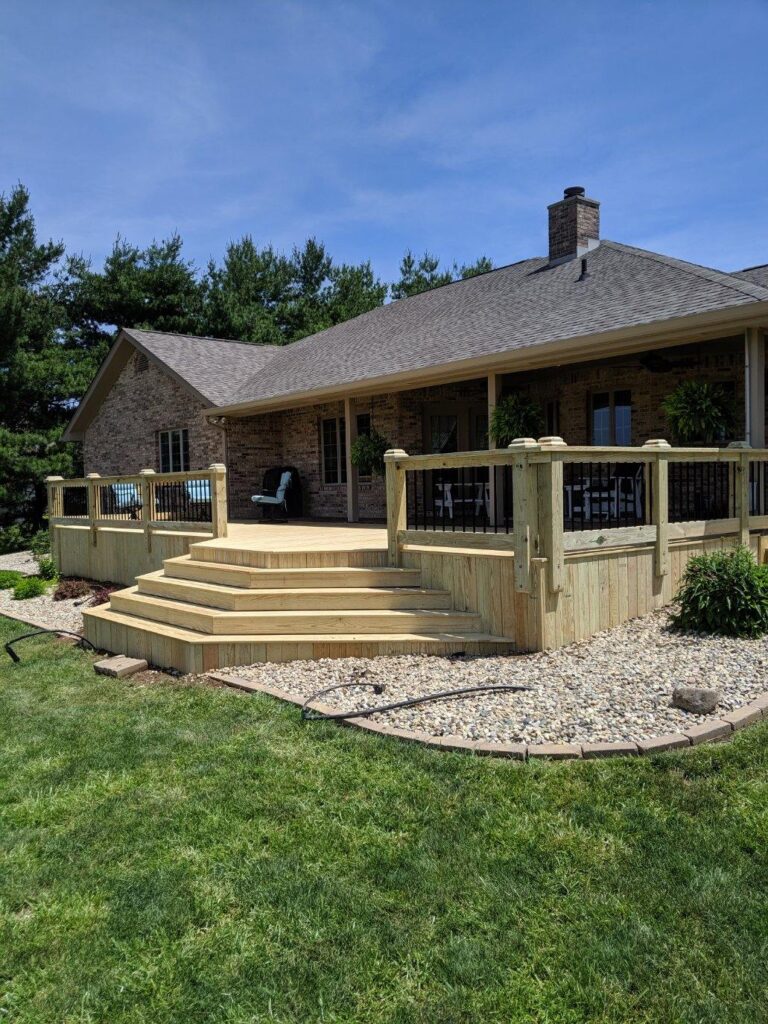A well-constructed wooden deck with steps leading up to a brick house, surrounded by lush green grass and landscaping.