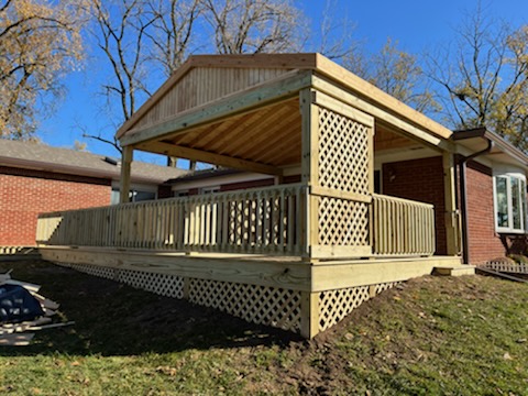 Newly built wooden deck with a gabled roof, featuring decorative lattice and railings, set against a backdrop of trees and a residential brick home.