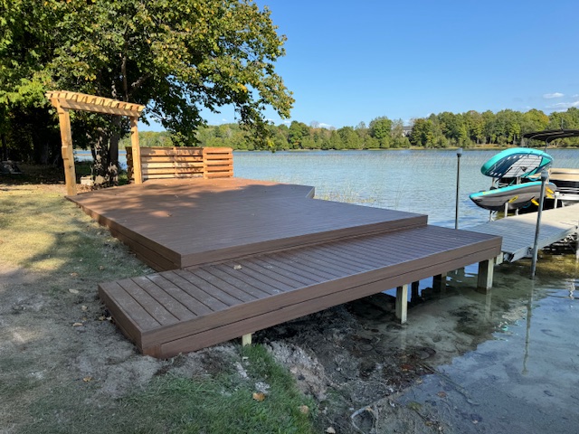 A newly constructed wooden deck by a lake featuring a pergola, with a boat dock and moored boat in the background.