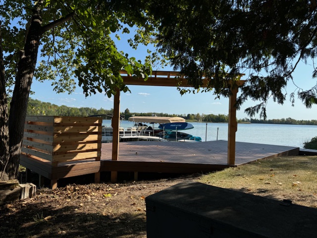 A lakeside deck with wooden railings and a pergola overlooking boats on the water, framed by green trees.