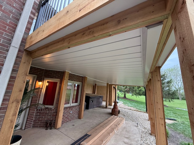 View of a covered outdoor patio featuring wooden beams, a stone path, and landscaping beyond.