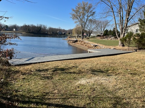 Scenic view of a serene lake with a wooden dock and grassy shoreline, surrounded by trees and residential homes.