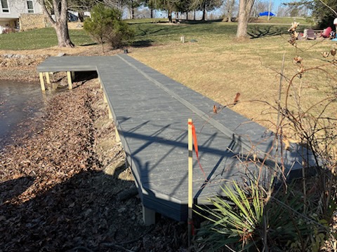 A wooden dock extends over a dry lake bed, surrounded by grass and trees, with a house visible in the background.