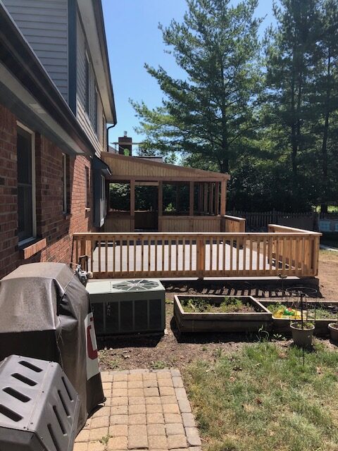 A well-lit backyard featuring a newly constructed wooden deck with a railing, adjacent to a brick house and a gas grill, surrounded by trees.