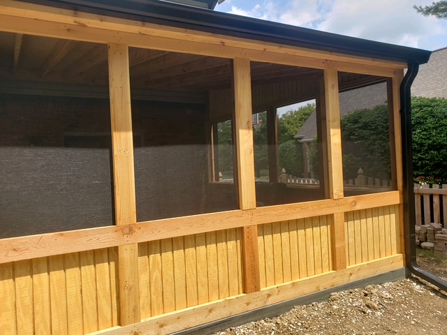 A wooden screened porch featuring a sturdy frame and mesh panels, with a light wood finish, surrounded by greenery and a neighboring house.