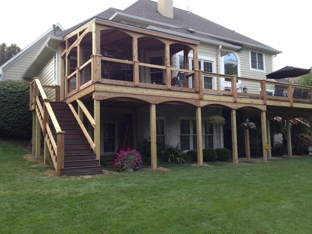 A two-story house featuring a wooden deck with stairs leading to the backyard, surrounded by lush green grass and flower pots.