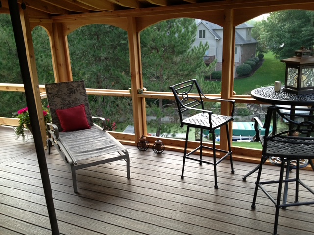 Screened porch with a chaise lounge, a bar stool set, and a table, surrounded by greenery and houses.