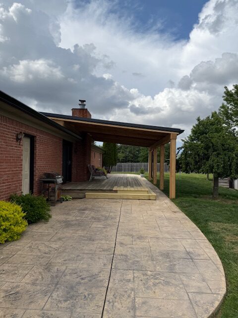 A view of a brick house featuring a wooden pergola and a patio with a paved path leading to a grassy backyard under a partly cloudy sky.