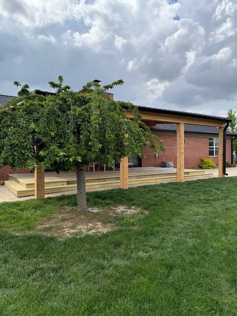 A wooden deck with posts is attached to a brick house, surrounded by green grass and a small tree under cloudy skies.