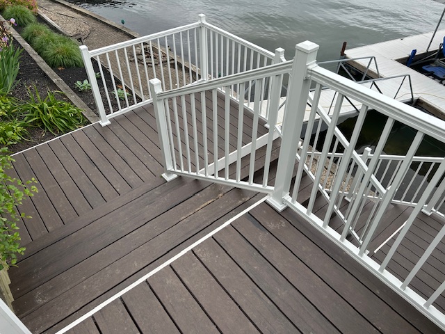 A wooden deck with a white railing leads down to a body of water, featuring a staircase and surrounding greenery.