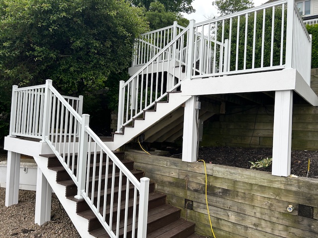 White wooden stairs with railings leading up to a deck, surrounded by greenery and wooden landscaping.