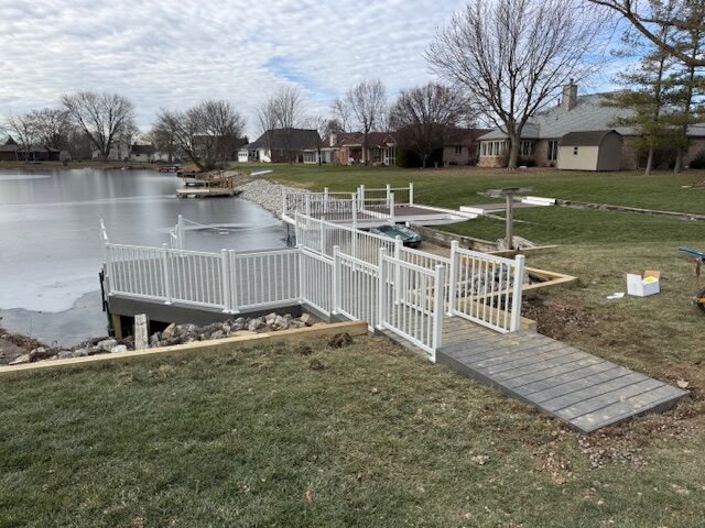 "View of a constructed dock area with white fencing and pathways leading to a frozen lake, surrounded by residential homes."