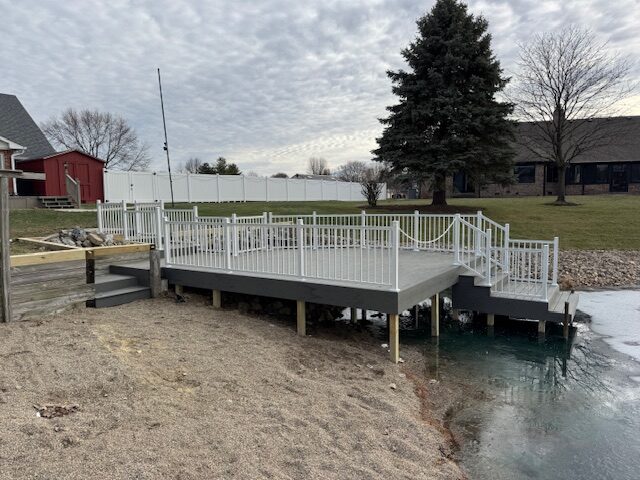 A gray dock with white railings extends over a calm body of water, surrounded by sandy bank and grass, under a cloudy sky.