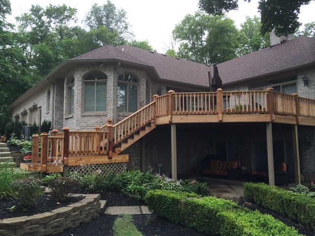 A modern brick house with a wooden deck and landscaped garden, surrounded by trees.