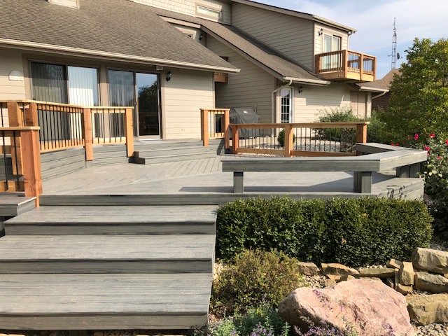 A spacious wooden deck with railings, surrounded by landscaping, leading to a two-story house.