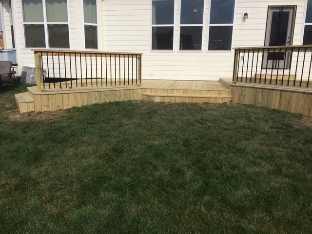 "Wooden deck with black railings, featuring steps leading to a grassy area and white house exterior."