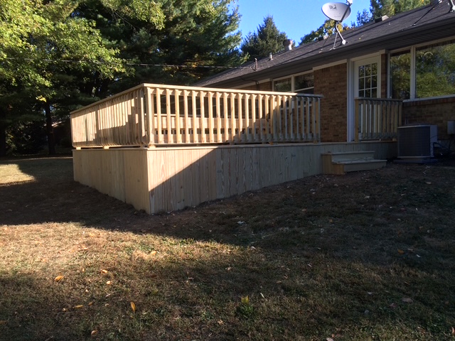 "Outdoor wooden deck extending from a brick house with an air conditioning unit, surrounded by grass and trees."