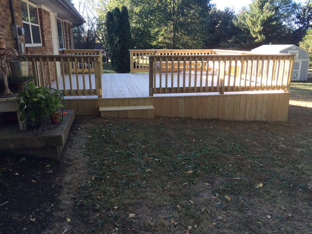 A spacious wooden deck with railings, built off the back of a house and surrounded by a yard with grass and landscaping.