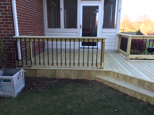 Wooden deck with a rail featuring black balusters, leading to a set of stairs, adjacent to a brick house.
