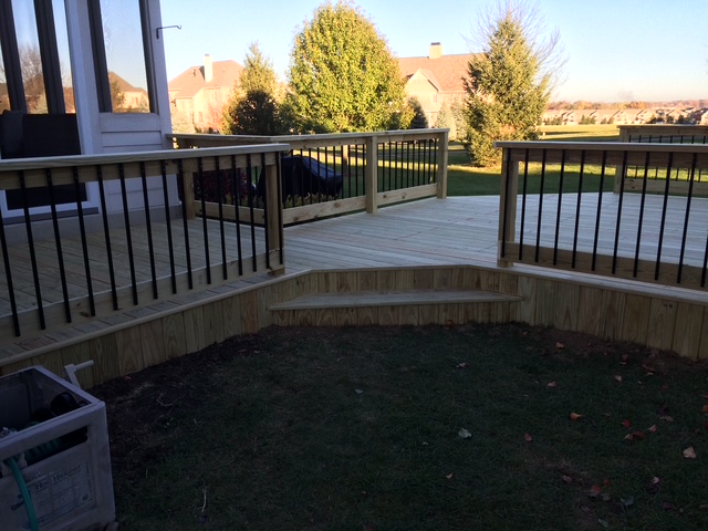 "Newly constructed wooden deck with black railings and steps leading to a grassy yard in a residential area."