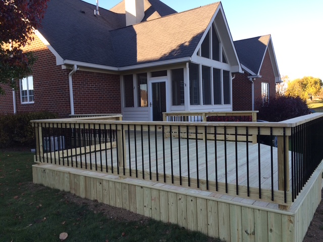 "New wooden deck with black railing attached to a brick house, featuring large windows and a well-maintained lawn."