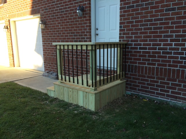 "Wooden entryway deck with black railings against a brick wall and a white door, featuring steps leading down to grass."