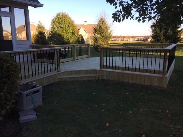 A newly constructed wooden deck with black railing, set in a spacious yard surrounded by greenery and residential homes in the background.