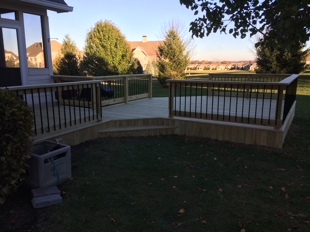 "New wooden deck with black railing in a residential backyard, surrounded by landscaped trees and grass."