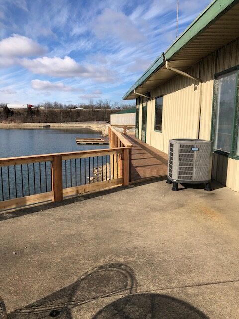 A view of a waterfront property featuring a wooden deck, air conditioning unit, and a river in the background under a partly cloudy sky.