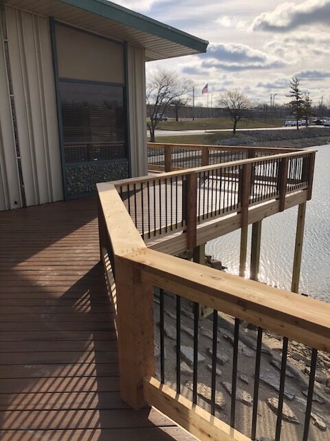 Wooden deck with a curved railing overlooking a body of water and a nearby building under a cloudy sky.