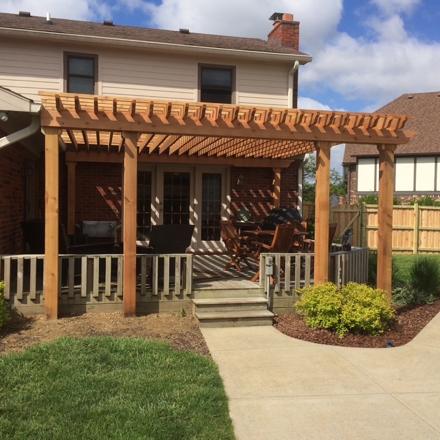 Wooden pergola over a patio area with seating, leading to a house with a well-maintained lawn and landscaped garden.